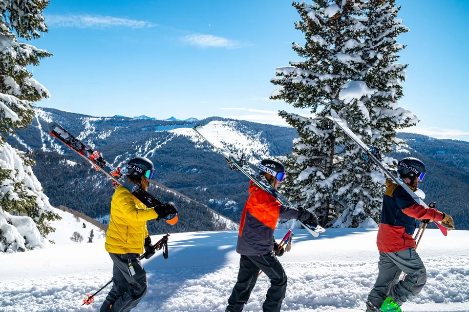 Wintersportler mit geschulterten Skiern vor verschneiten Tannen und Bergpanorama